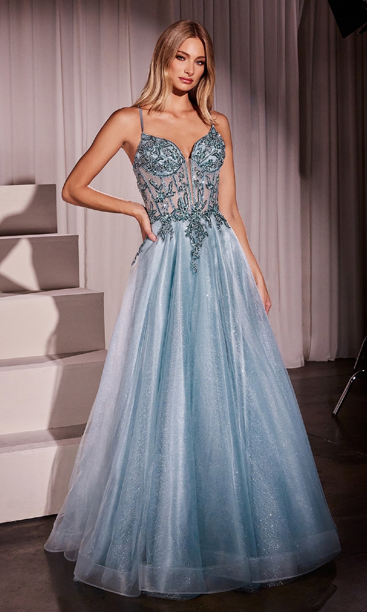 A woman stands indoors wearing the Sheer-Corset Long Prom Dress Ladivine CC0881, featuring a light blue A-line silhouette with a beaded embroidered bodice and a voluminous tulle skirt.